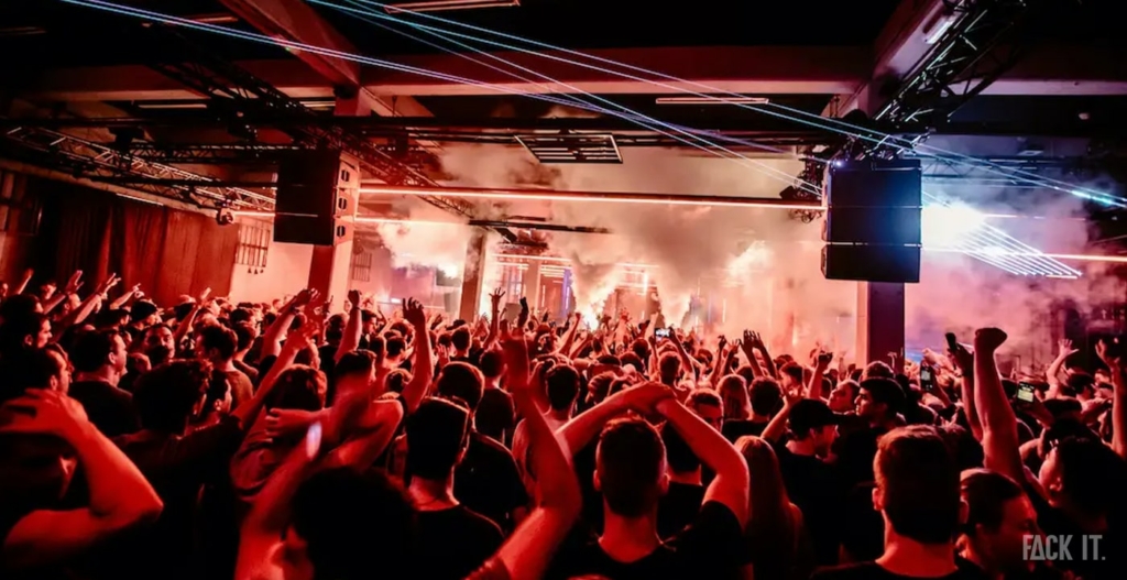 A vast, gritty warehouse with exposed brick, steel beams, and a dark, electrifying atmosphere filled with a crowd dancing to thumping techno beats.