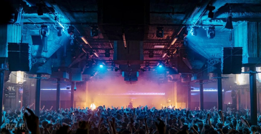 A massive, industrial venue with towering ceilings, thick concrete walls, and a crowd energized by powerful techno beats, all bathed in dynamic lighting.