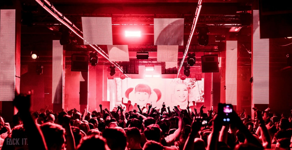 A massive industrial warehouse with towering ceilings, exposed steel beams, and a sea of ravers immersed in deep, pounding techno.