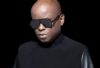 Juan Atkins, wearing glasses and posing against a dark background, gazes thoughtfully away from the camera, embodying his role as the pioneer of techno music.